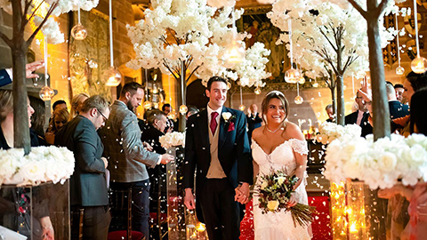 A man wearing a suit and a woman wearing a white wedding dress walking through a brightly lit venue surrounded by white flowers with other people in the background